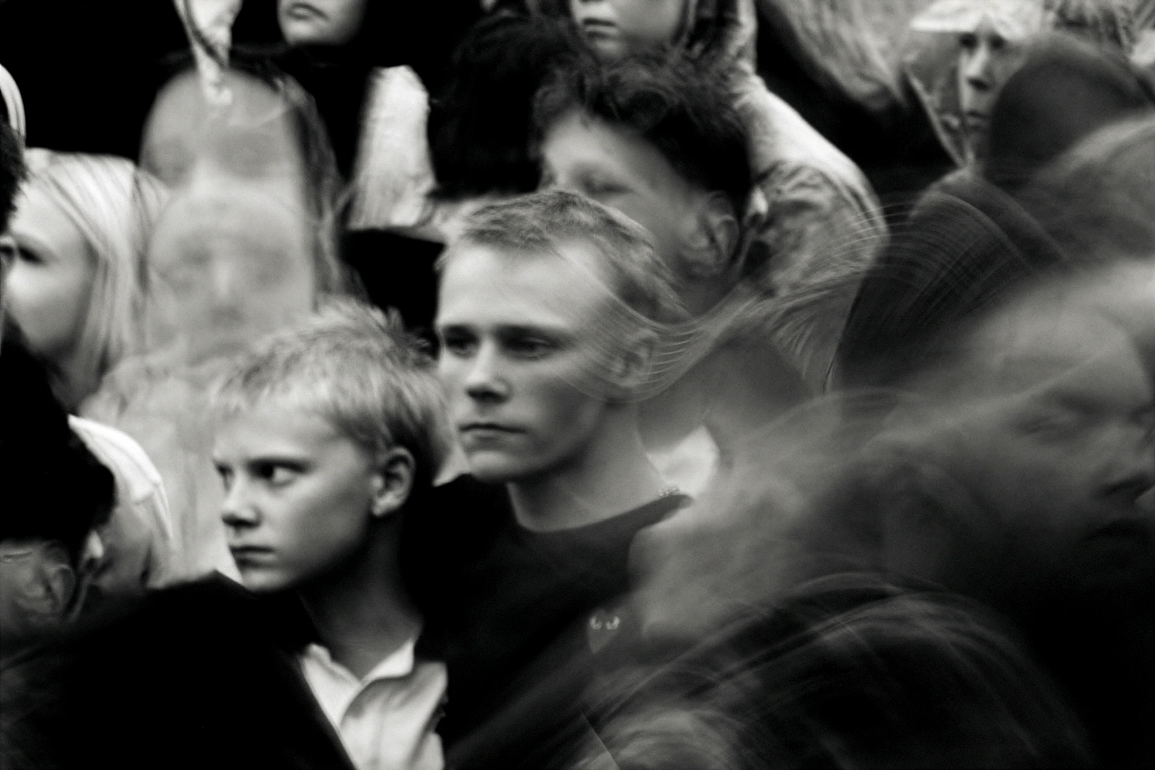Two young people standing in a crowd with motion blurred reflections surrounding them. One boy looks towards the left while another young man stands behind him.