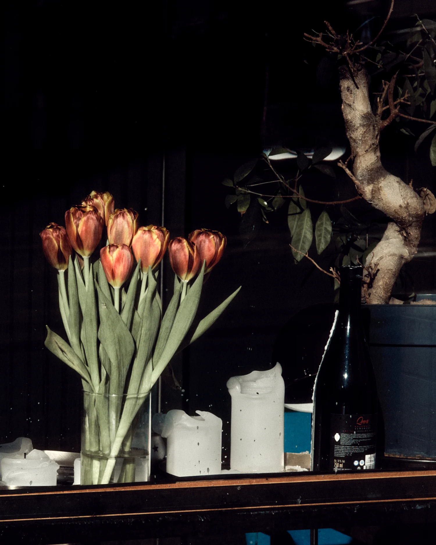 A bouquet of red and orange tulips in a clear glass vase displayed in a window in Helsinki. White candles, a dark wine bottle, and a small potted plant sit alongside the flowers against a dark interior background.
