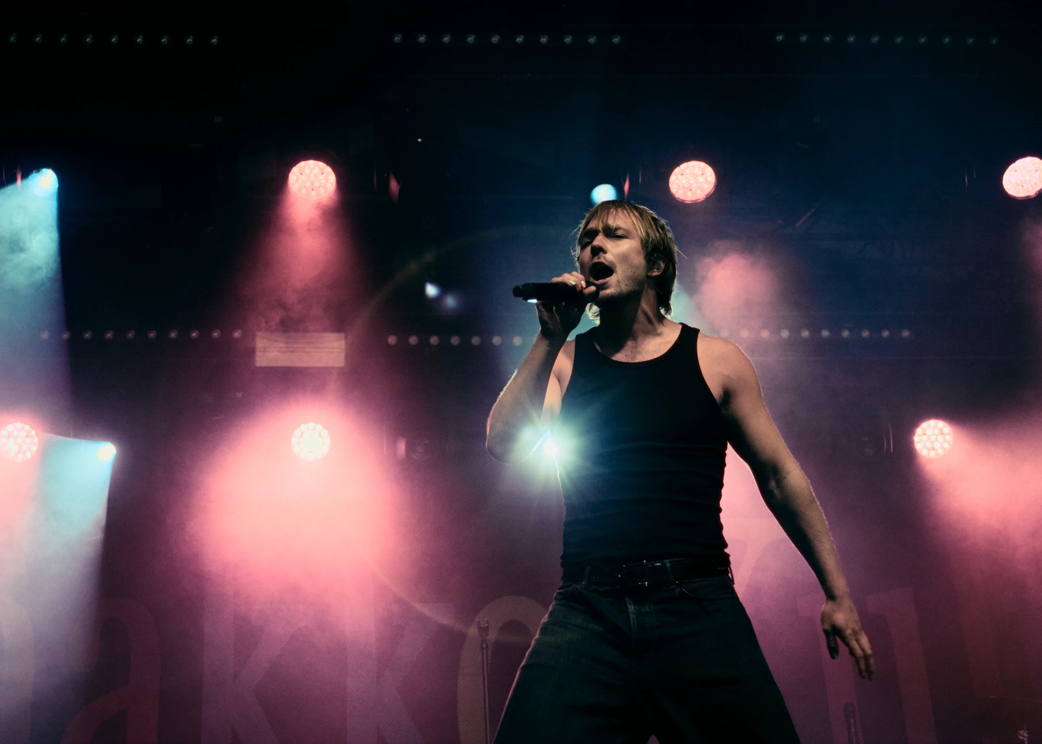 A male singer performing on stage with a microphone. He is wearing a black tank top and is illuminated by red and white stage lights at Qstock 2026.