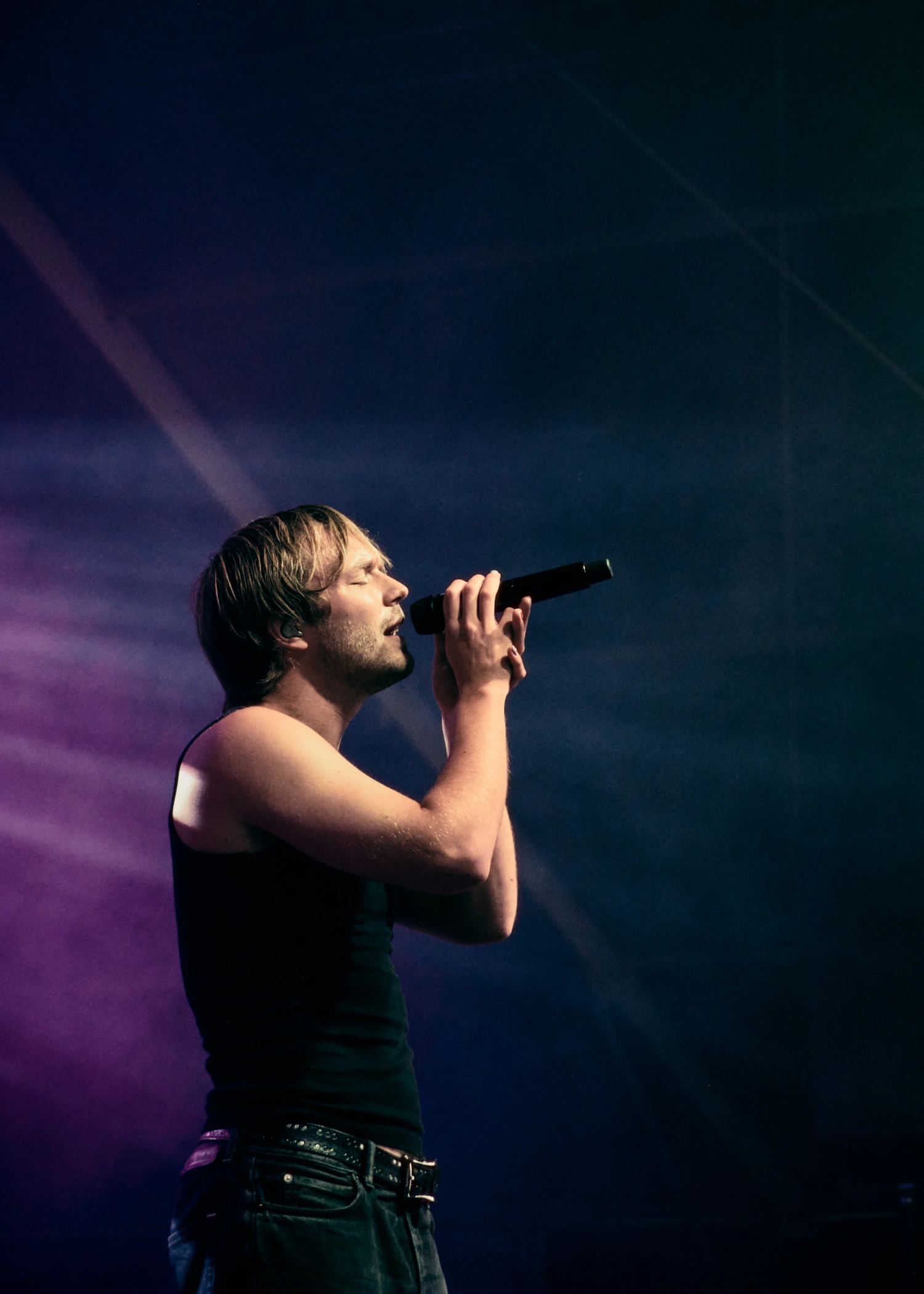 A side profile of a male singer on stage holding a microphone with both hands. He is wearing a black tank top and dark jeans.