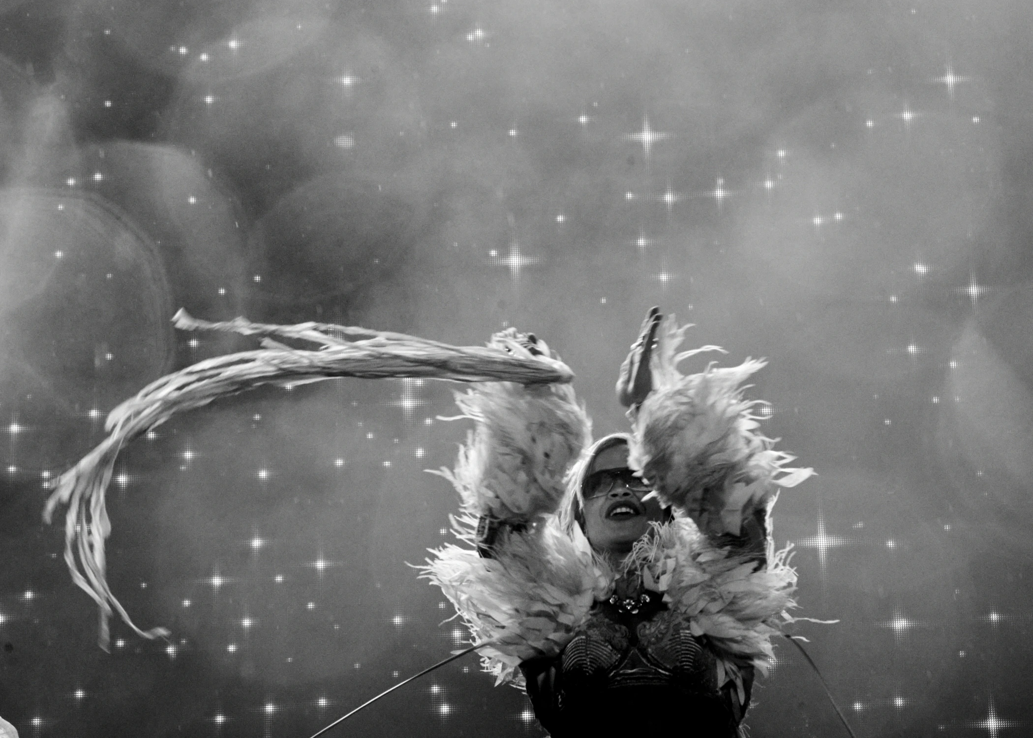 A black and white view of a performer in a feathery outfit and sunglasses holding long tassels up. The background has a starry, glittery effect.