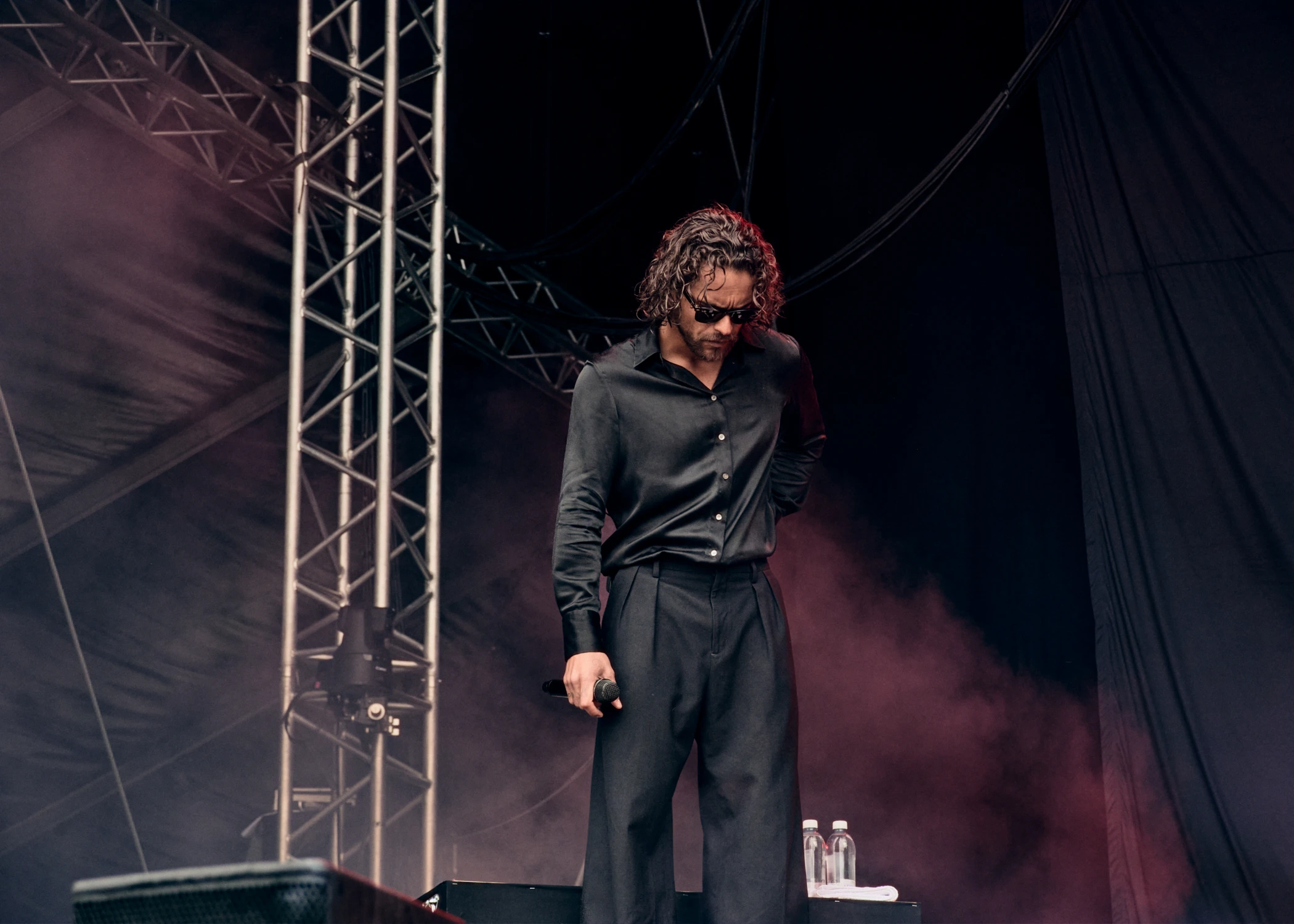 A male singer with dark curly hair and sunglasses performing on stage. He is dressed in a black button-up shirt and black trousers.