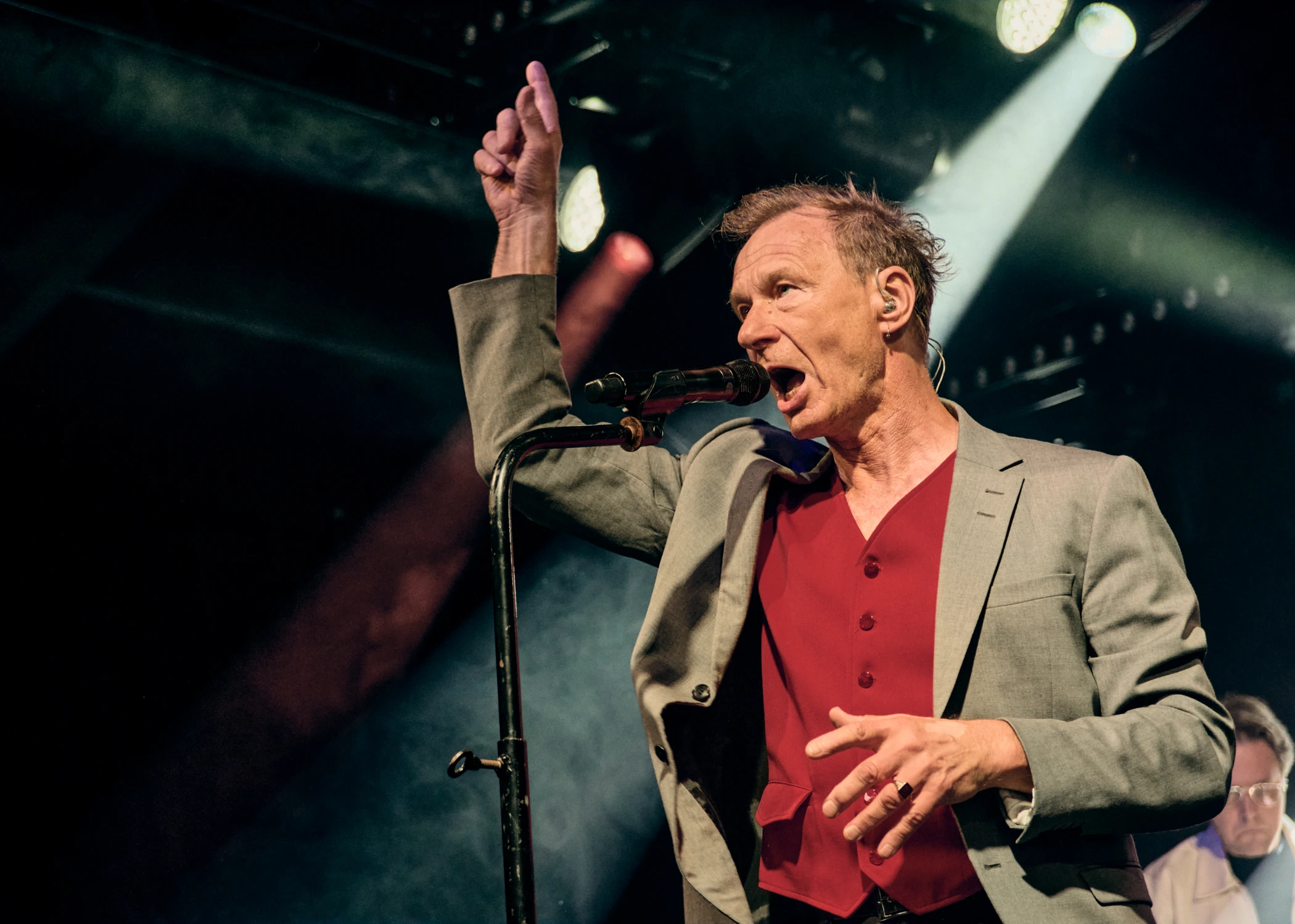 A male singer performing on stage, singing into a microphone with his right arm raised. He is wearing a grey jacket over a red vest under bright stage lights.