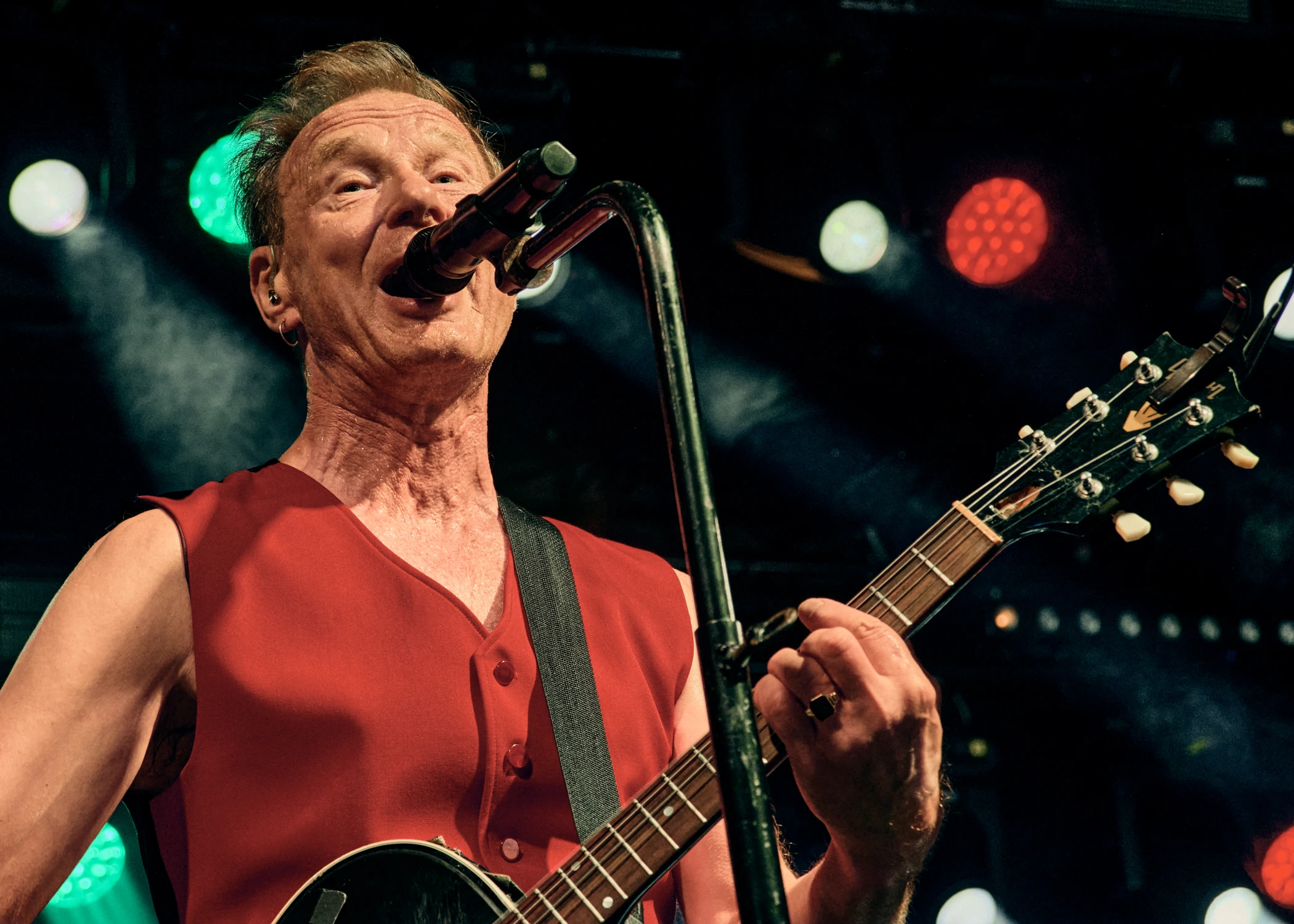 A male singer playing an electric guitar and singing into a microphone. He is wearing a red sleeveless vest with green and red stage lights behind him.