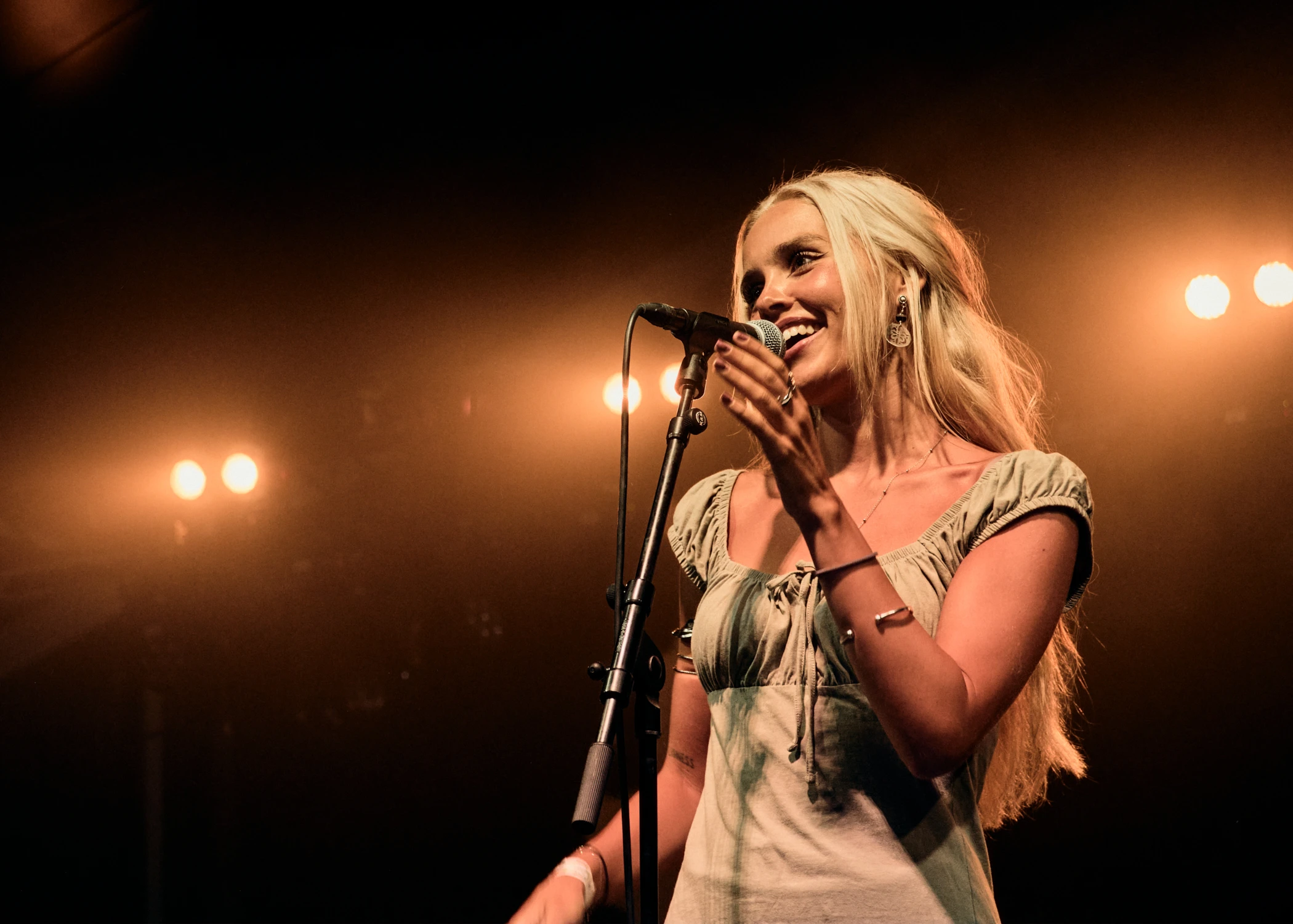 A blonde female singer smiling and speaking into a microphone on stage. She is wearing a light-colored dress and large earrings under warm stage lights.