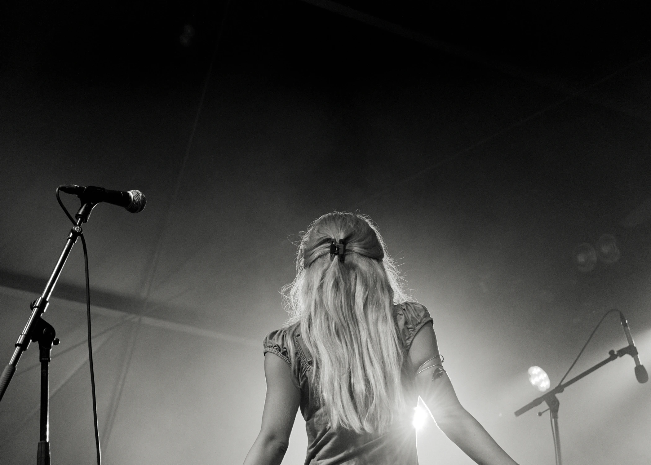 A black and white view from behind a female singer with long blonde hair on stage. She is facing bright stage lights with a microphone stand to her left.