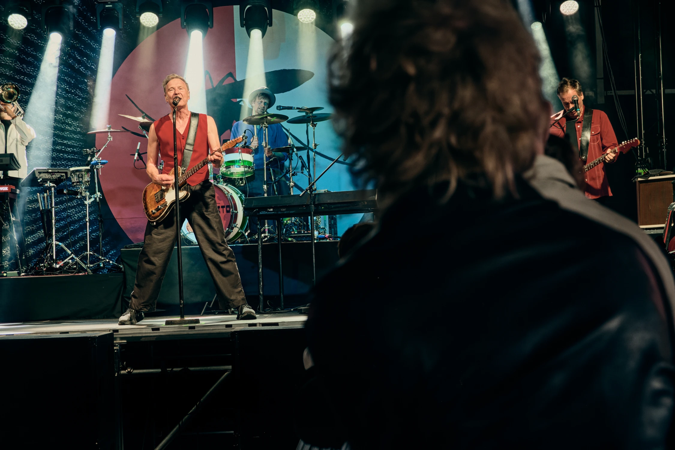 A male singer playing an electric guitar and singing on stage. He is wearing a red vest and black trousers, with a drummer and another guitarist visible in the background behind him. A blurred head of an audience member is in the foreground.