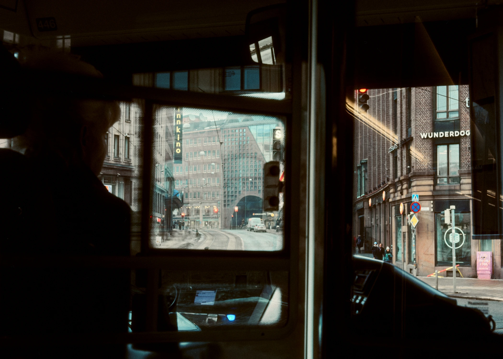 A view from inside a tram looking out through the windows onto a street in Helsinki. The front window frames tram tracks leading towards a tunnel between city buildings, while the tram driver is seen in silhouette on the left.
