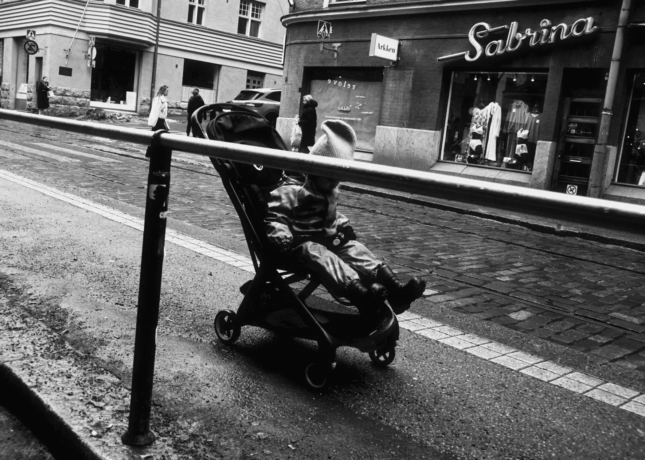 A black and white street view of a small child sitting in a stroller on a wet cobblestone street in Helsinki. A metal railing runs horizontally across the foreground, while a shop named Sabrina is visible in the background.