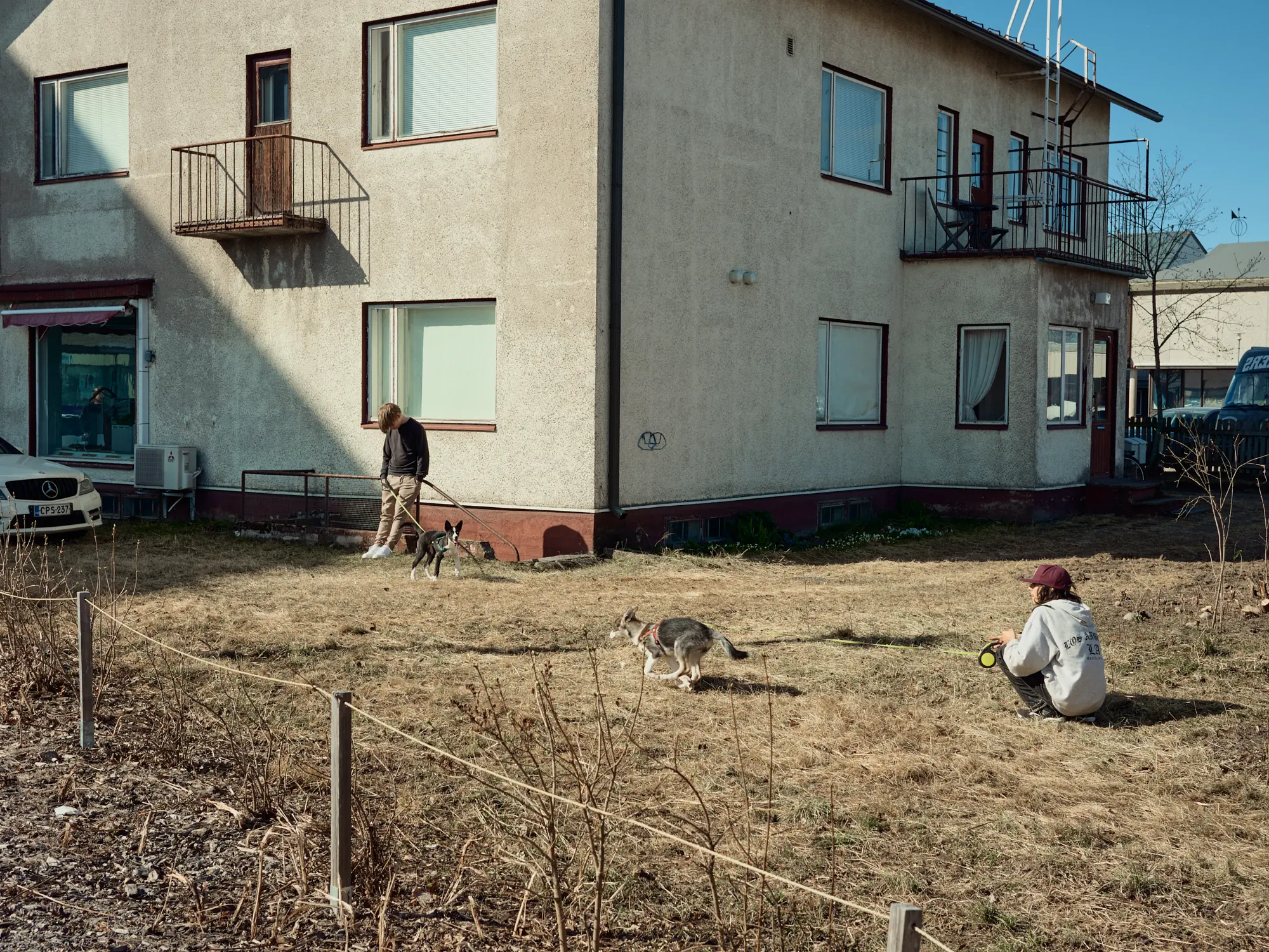 Two young people walking and playing with their dogs on the lawn of an apartment building in bright spring sunlight in Ylivieska, Finland.