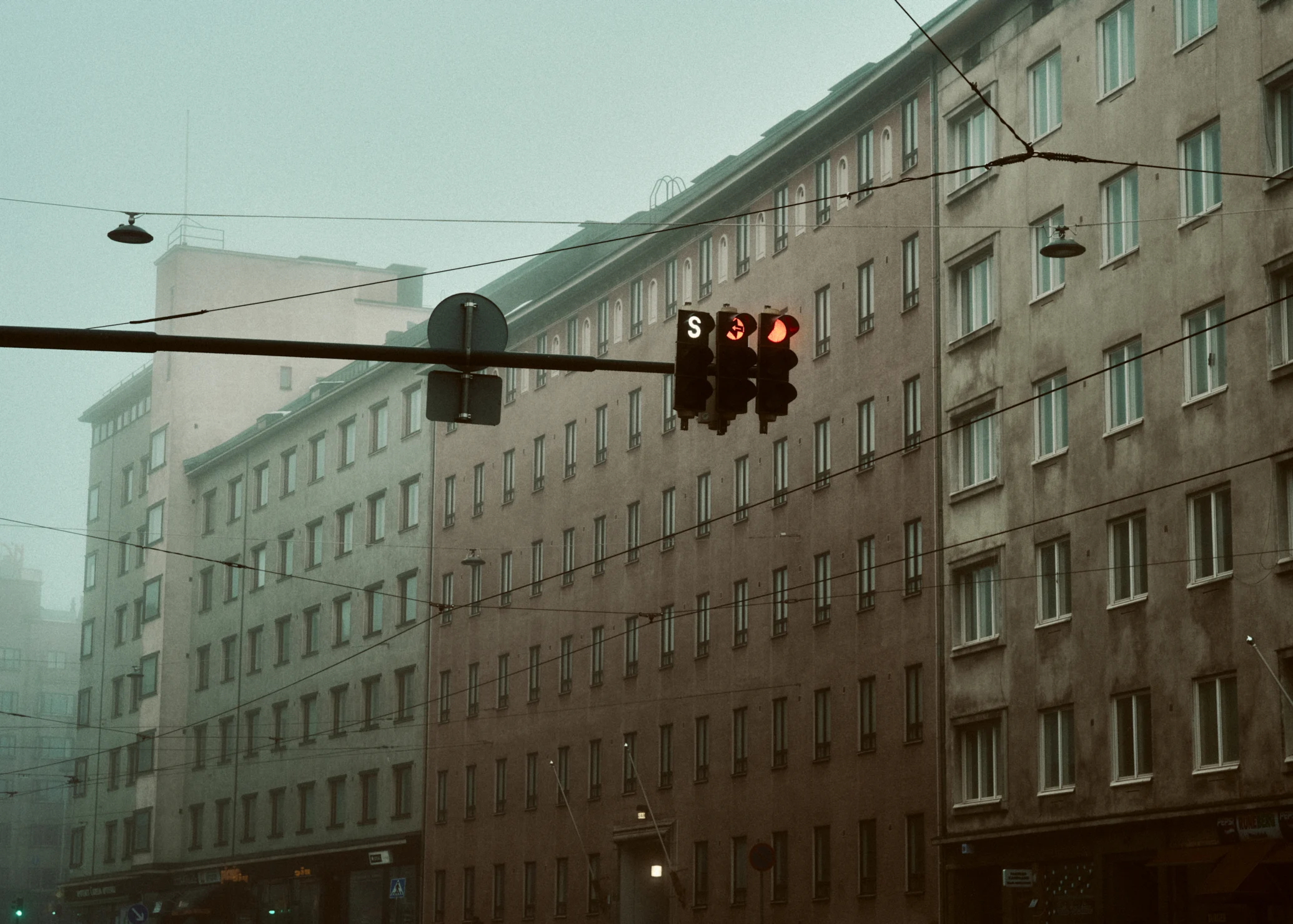 Traffic lights showing red and a white "S" tram signal on a street in Helsinki. Multi-story apartment buildings line the street behind them on a hazy day with overhead tram wires crossing the sky.