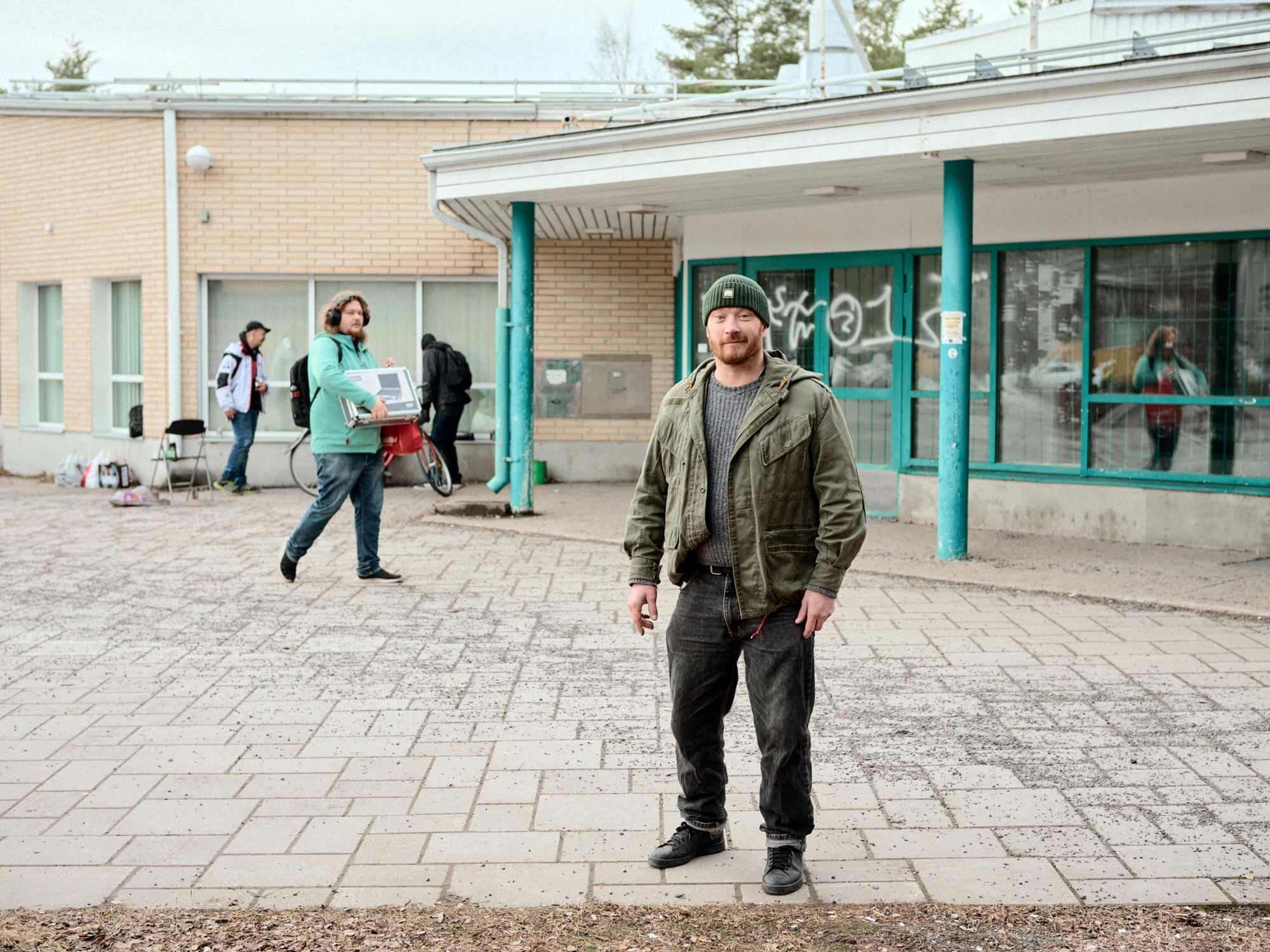 A red-bearded man in a green beanie and olive jacket standing on a paved square in front of a weathered brick building with teal pillars and graffiti in Kaijonharju, Oulu. Other people pass in the background.