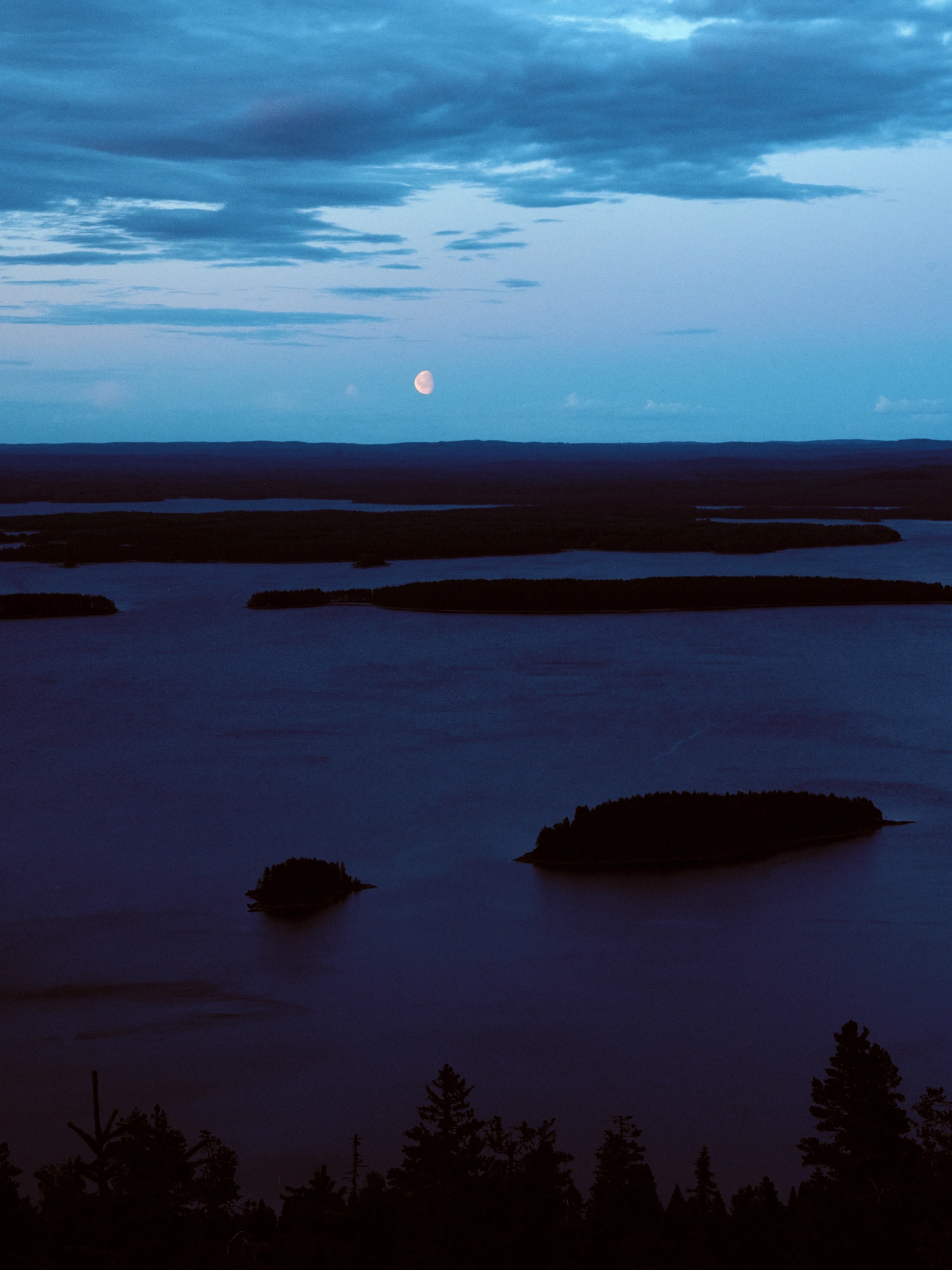 A tranquil twilight landscape looking out over Lake Pielinen from Koli, with silhouetted forested islands on dark blue water under an evening sky with the moon.