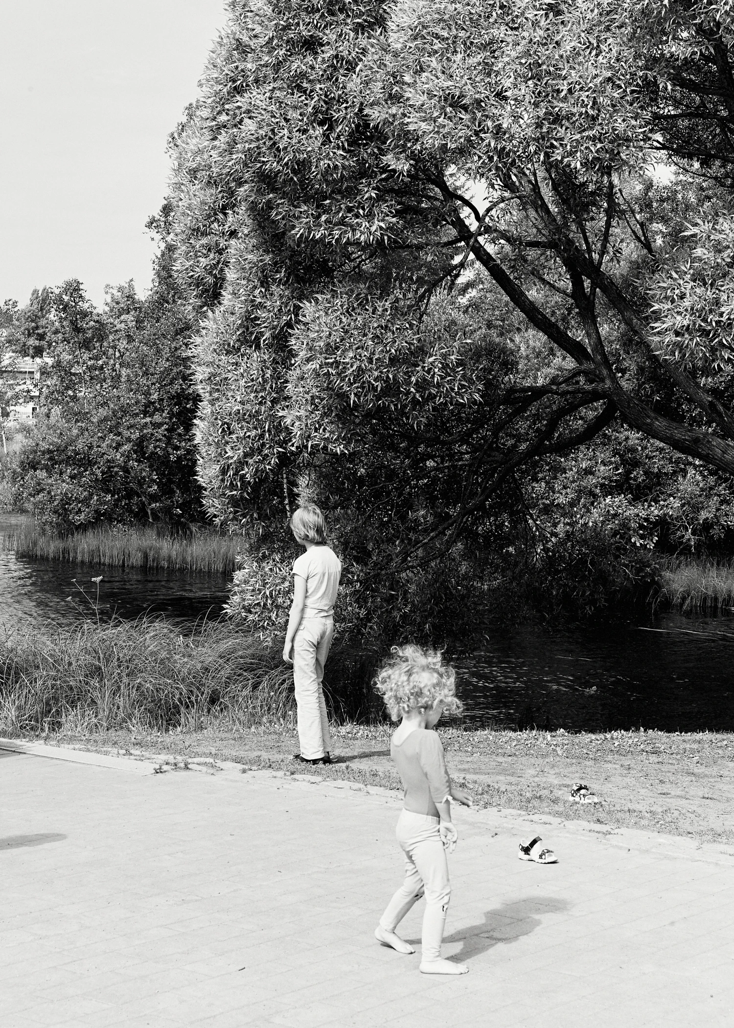 Two children in a park near a body of water under a large leafy tree. In the foreground, a barefoot toddler with curly hair walks on a paved path, with their sandals left on the grass nearby. Further back, an older child stands on the grassy bank looking towards the water.