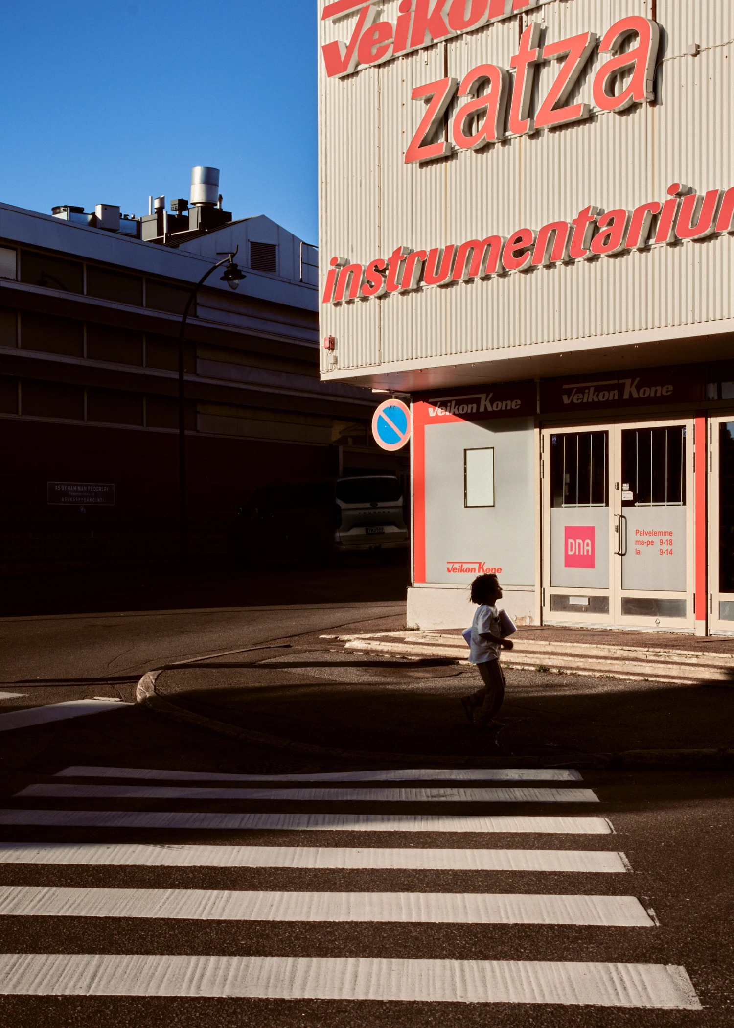 A small child walks near a pedestrian crosswalk in front of a white commercial building with large red signs. Sunlight illuminates the building's corner, while the street behind it is in deep shadow.