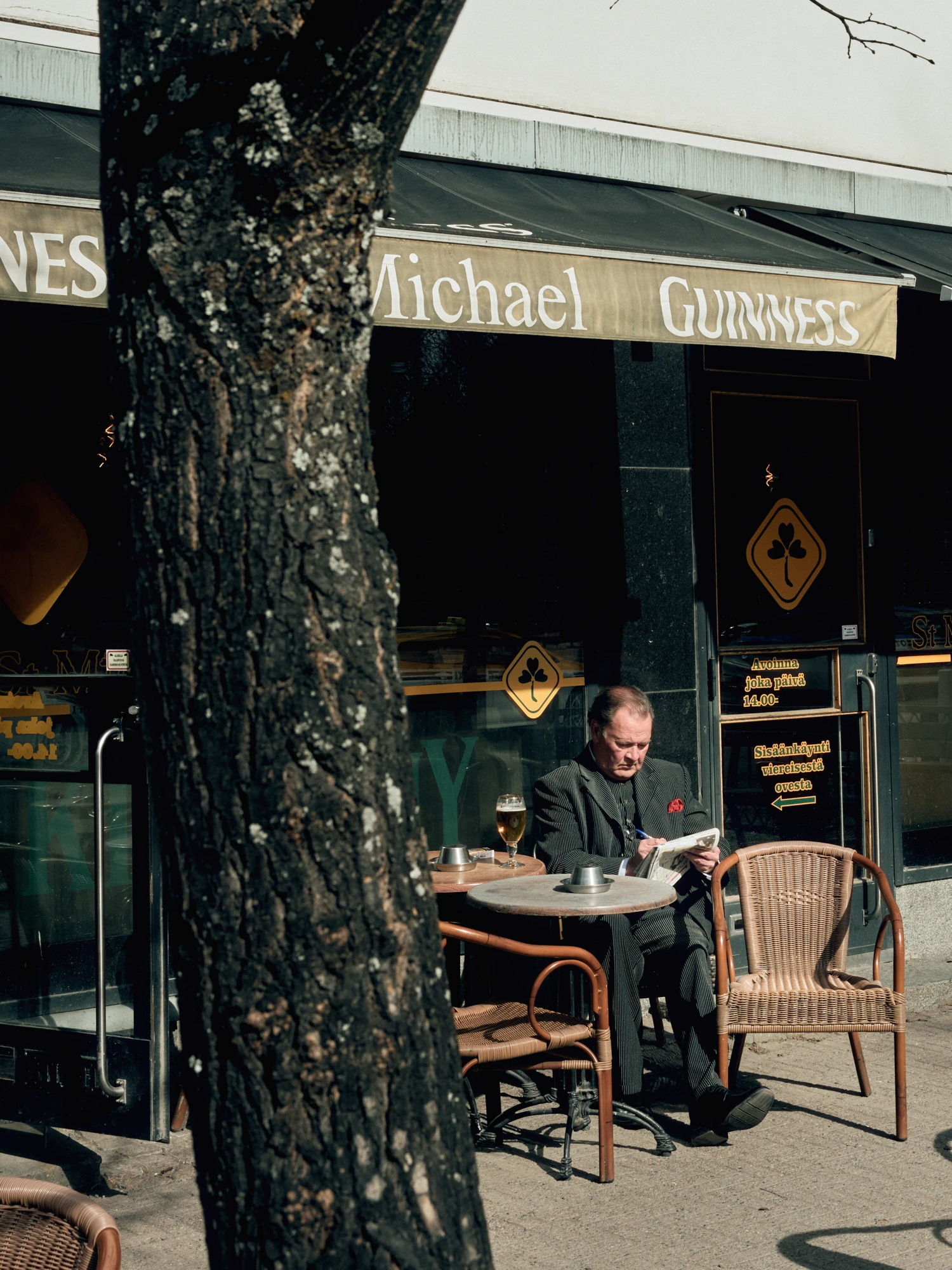 A man in a dark suit sits alone at an outdoor table outside Michael Guinness pub in Oulu, reading a newspaper. A beer glass and an ashtray are on the table. A thick tree trunk fills the foreground.