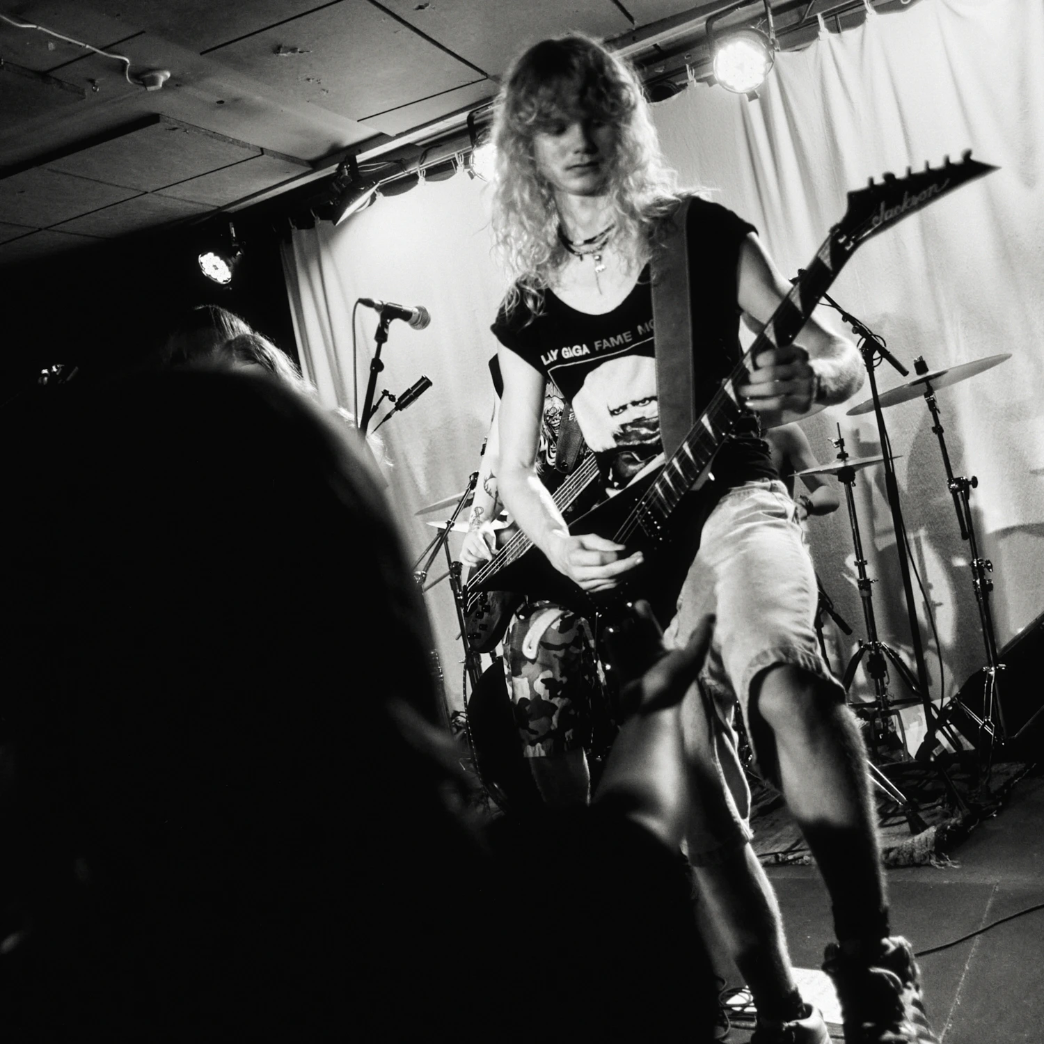 A black and white gig photo of a band performing live on stage. The musicians are captured in action with stage lighting illuminating them.