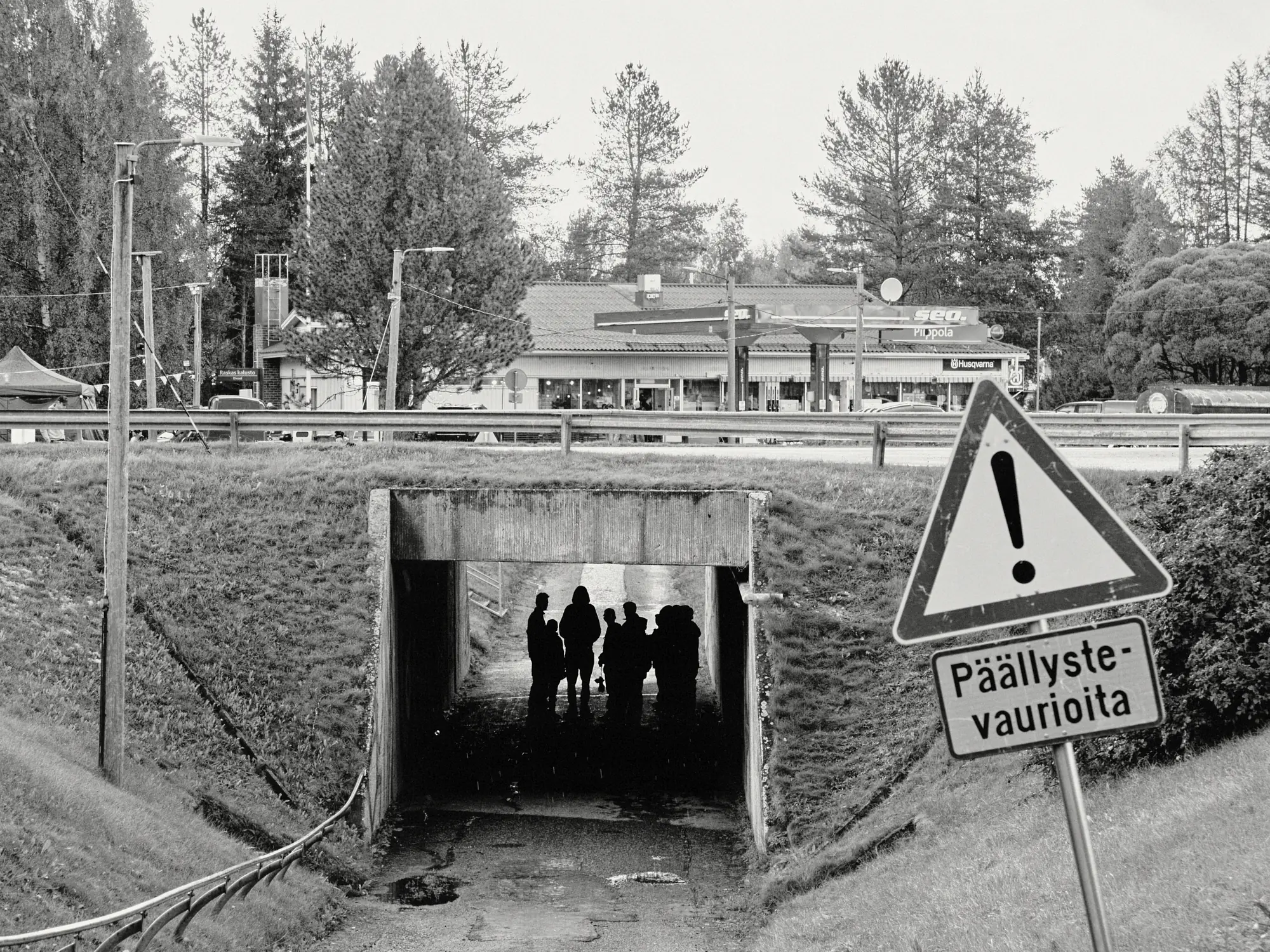 Mustavalkoinen teos, jossa etualalla seisoo kolmionmuotoinen varoitusmerkki tekstillä 'Päällystevaurioita'. Betonisessa alikulkutunnelissa erottuu ryhmä nuoria tummina siluetteina varjossa, ja taustalla kohoaa maaseututaajaman huoltoasema mäntyjen reunustamana Piippolassa.