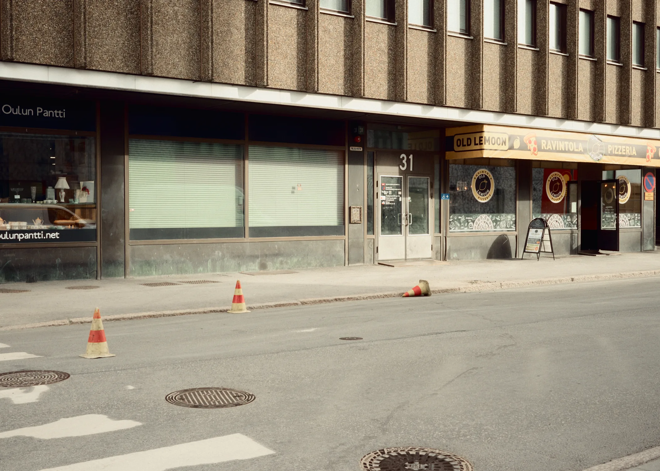 An urban street scene in Oulu featuring a brown concrete building facade, Oulun Pantti pawn shop, a yellow awning for Old Le Moon restaurant, and orange traffic cones on the asphalt.