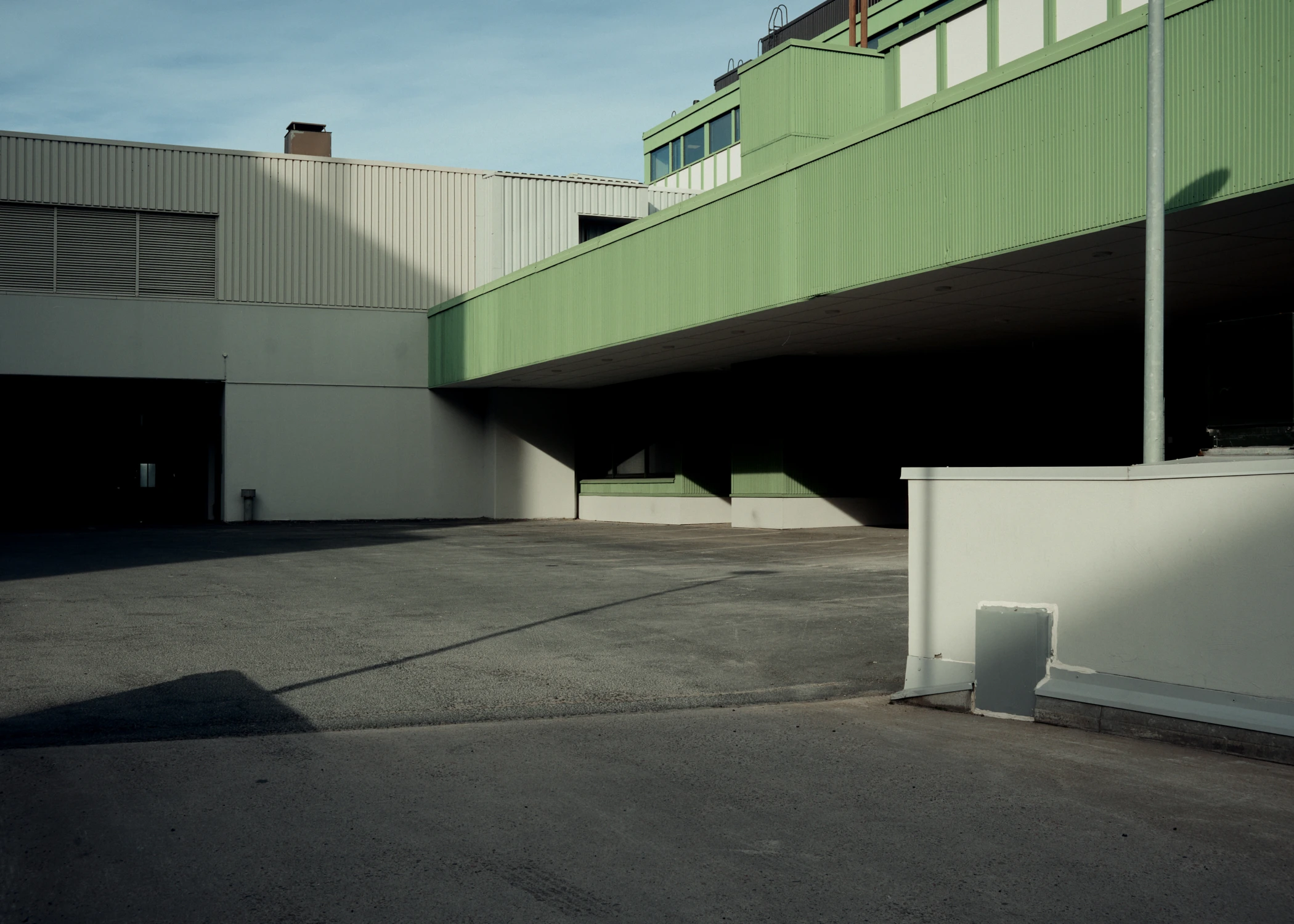 An empty asphalt yard surrounded by industrial buildings. A green corrugated overhang casts deep, diagonal shadows across the ground in bright daylight. Oulu, 2026.